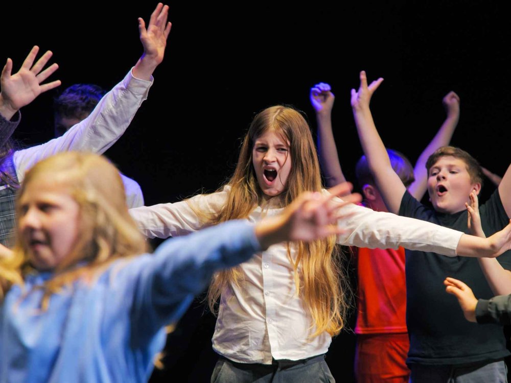 A group of children perform energetically on stage with their arms raised during a theatre workshop or performance. In the centre, a girl with long hair and a white shirt sings or shouts expressively while other children around her strike dramatic poses under colourful stage lighting against a dark background.