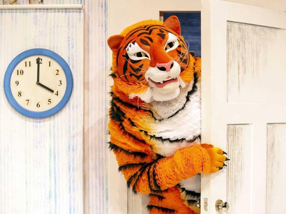 A performer in an orange tiger costume peeks cheerfully around a white door with a blue clock on the wall behind.