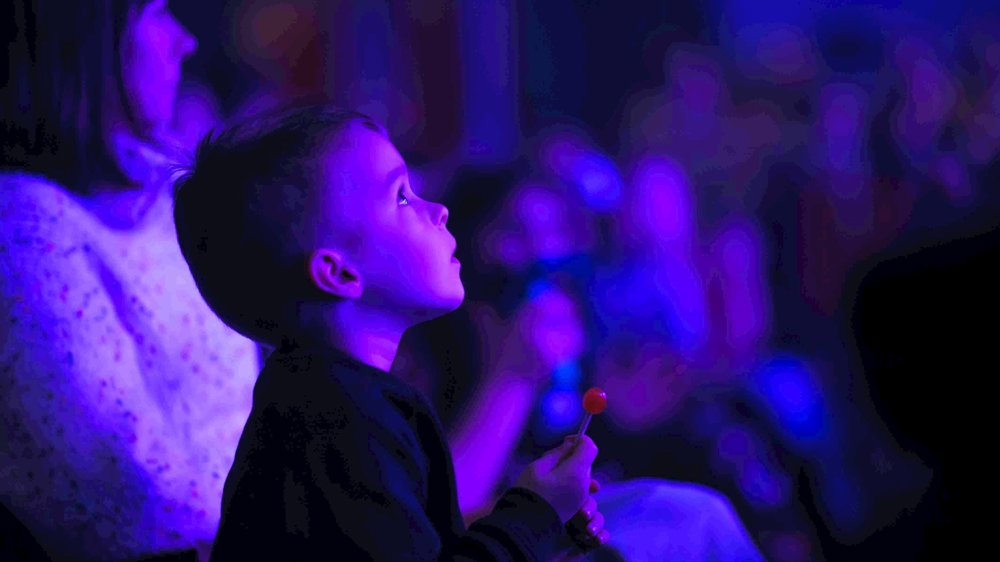 A young boy sitting in a theatre audience, holding a red lollipop and looking upwards with a sense of wonder.