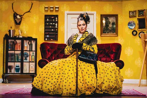 A character wearing a big, yellow and black spooted dress is sat on a red velvet chair. In their black gloved hands, they rest on a walking stick. 