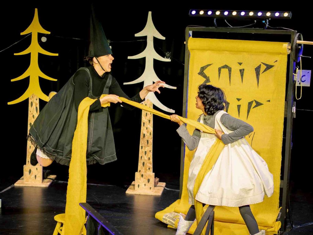 Rapunzel and the witch engage in a tug-of-war with Rapunzel's long yellow hair. Surrounded by stylised woodland scenery and a tower set, the pair act out a dramatic moment from the story.
