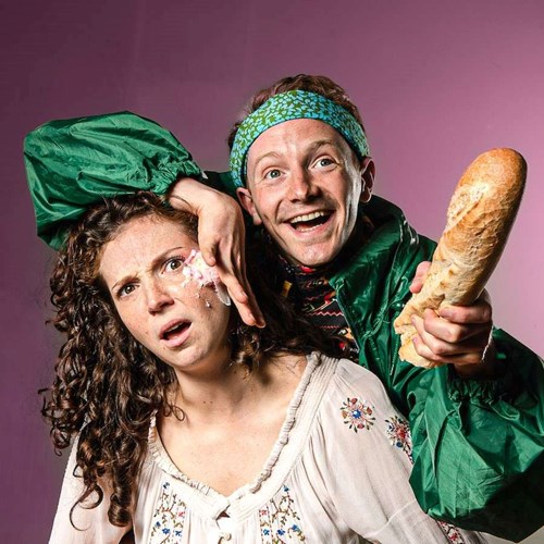 Two actors from Slapstick Picnic dressed as Wendy and Peter Pan. Peter is smiling, wearing a green bandana and a green coat. He is holding a baguette in one hand and smushing a cake into Wendy's cheek with the other. Wendy looks shocked. 