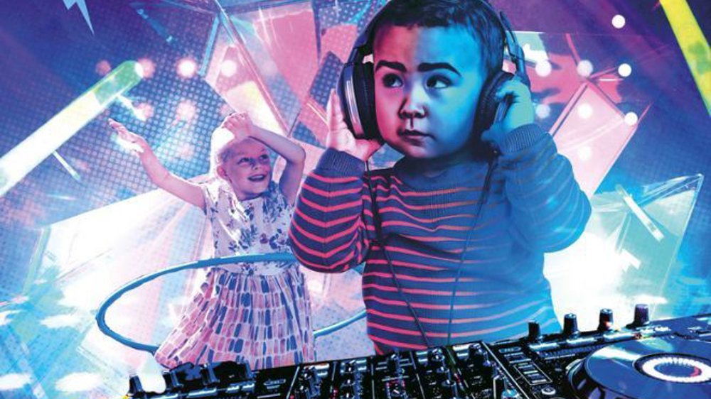 The image features a playful scene of young children in a disco-like environment. In the foreground, a toddler is wearing oversized headphones, standing at a professional DJ deck, as if they are mixing music. The child, dressed in a striped sweater, looks focused and serious while interacting with the equipment. In the background, another child is joyfully dancing, wearing a patterned dress, with arms raised and a hula hoop around their waist. 