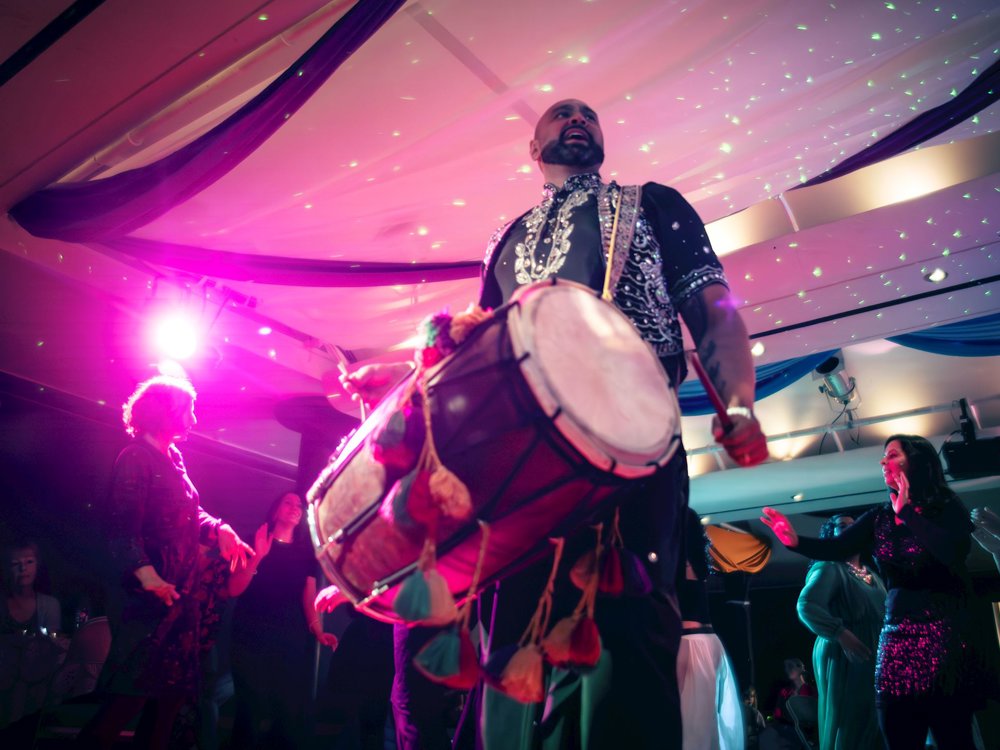 The image captures a vibrant and energetic Bhangra performance in an indoor setting, with colourful lighting creating a festive atmosphere. At the centre, a male performer wearing a traditional, ornate outfit is playing a large dhol drum, with tassels hanging from its sides. His stance is strong and commanding, and the bright stage lights cast a pink and purple glow over him and the room. Surrounding him, people are joyfully dancing, clapping, and moving to the rhythm of the music.