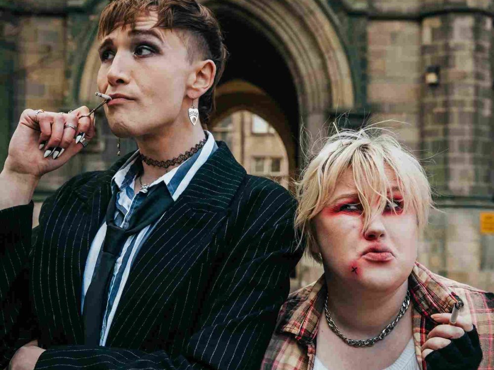 Two people stand closely outside a gothic archway. One lights a cigarette while the other leans in, both dressed in punk-inspired clothing with bold makeup and accessories.