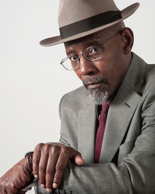 Linton Kwesi Johnson, writer and poet, poses wearing a beige hat, glasses and a grey suit with a red tie. 