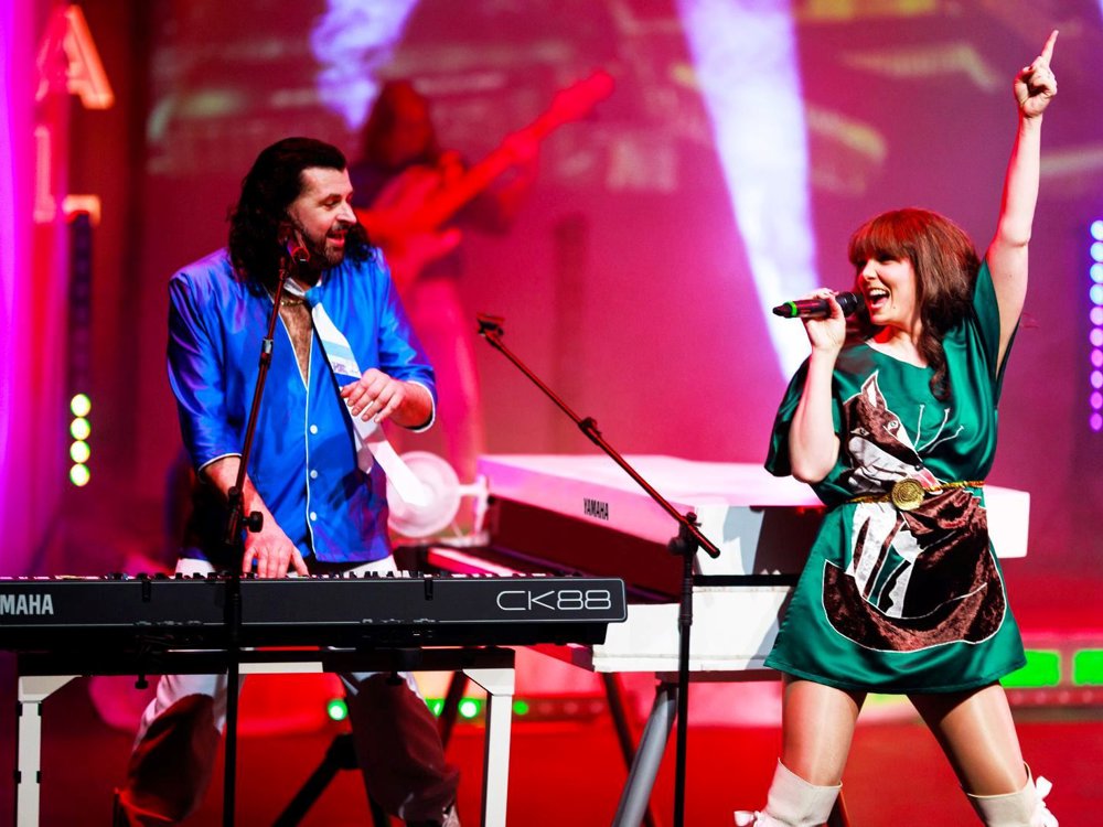 A singer in a green costume raises her arm as she performs beside the keyboard player, recreating an iconic ABBA performance with colourful stage lighting.