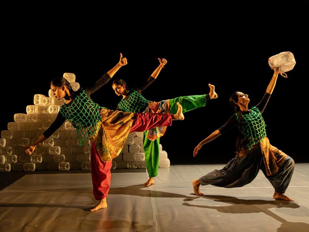 Three dancers perform on a dark stage, balancing in expressive poses while holding plastic containers. They wear colourful costumes with green netting overlays, with stacks of plastic containers forming part of the set behind them.