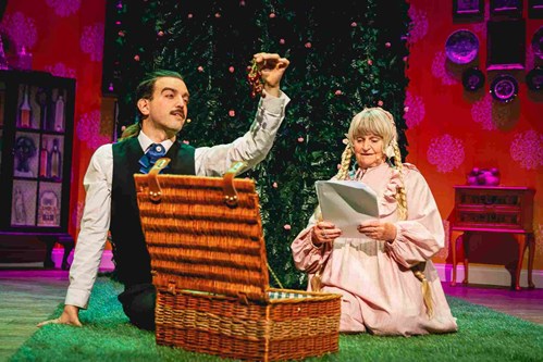 From a picnic basket inbetween two characters who are sat on the floor, someone holds a bunch of grapes. The character is wearing a black waist coat and blue tie. Another character is wearing a long, blond, plaited wig. They are reading from a script. 