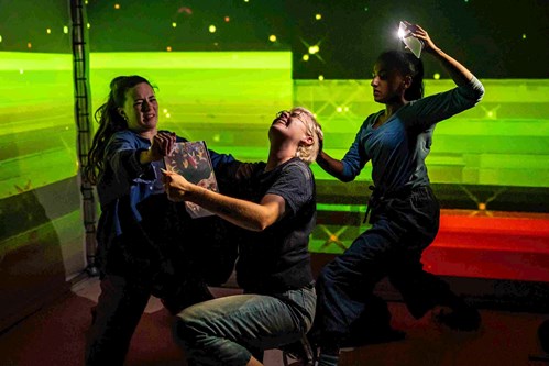 In a darkened space with a bright green light projected behind, are three young people. In the middle, is a young person on their knees. Their head is pulled back by another young person holding up their phone with the torch on. As they are on their knees, they are gripping a book. Another young person in front of them tries to pull the book from their hand. 