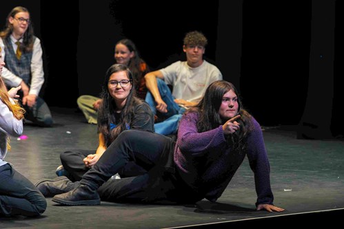 An older Jack performs on stage during a youth theatre production, confidently pointing while delivering a line. He wears a purple jumper and black trousers, surrounded by fellow cast members sitting on the stage in the background.