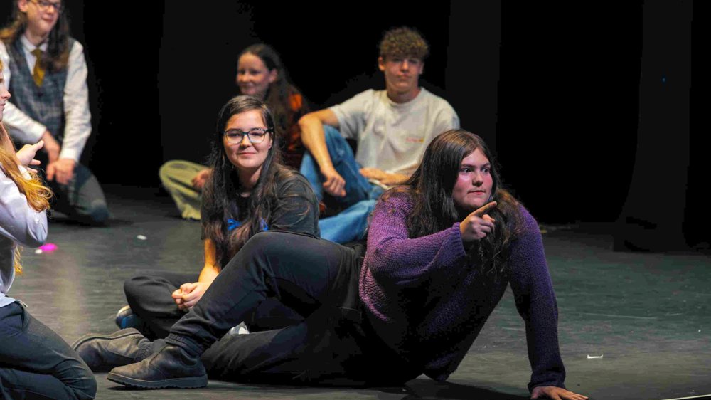 An older Jack performs on stage during a youth theatre production, confidently pointing while delivering a line. He wears a purple jumper and black trousers, surrounded by fellow cast members sitting on the stage in the background.