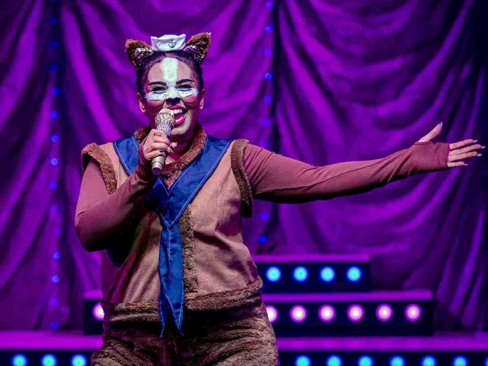  A performer dressed as a lively animal character, possibly a cat or dog, sings into a glittery microphone. The costume features brown fur-like fabric, a blue scarf-like accessory, and a headband with fuzzy animal ears. The performer's face is painted with whiskers and a white muzzle, adding to the character's playful look.