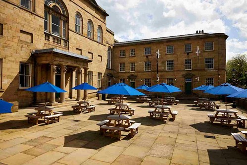 A courtyard with multiple round, wooden benches that each have blue umbrellas in. 