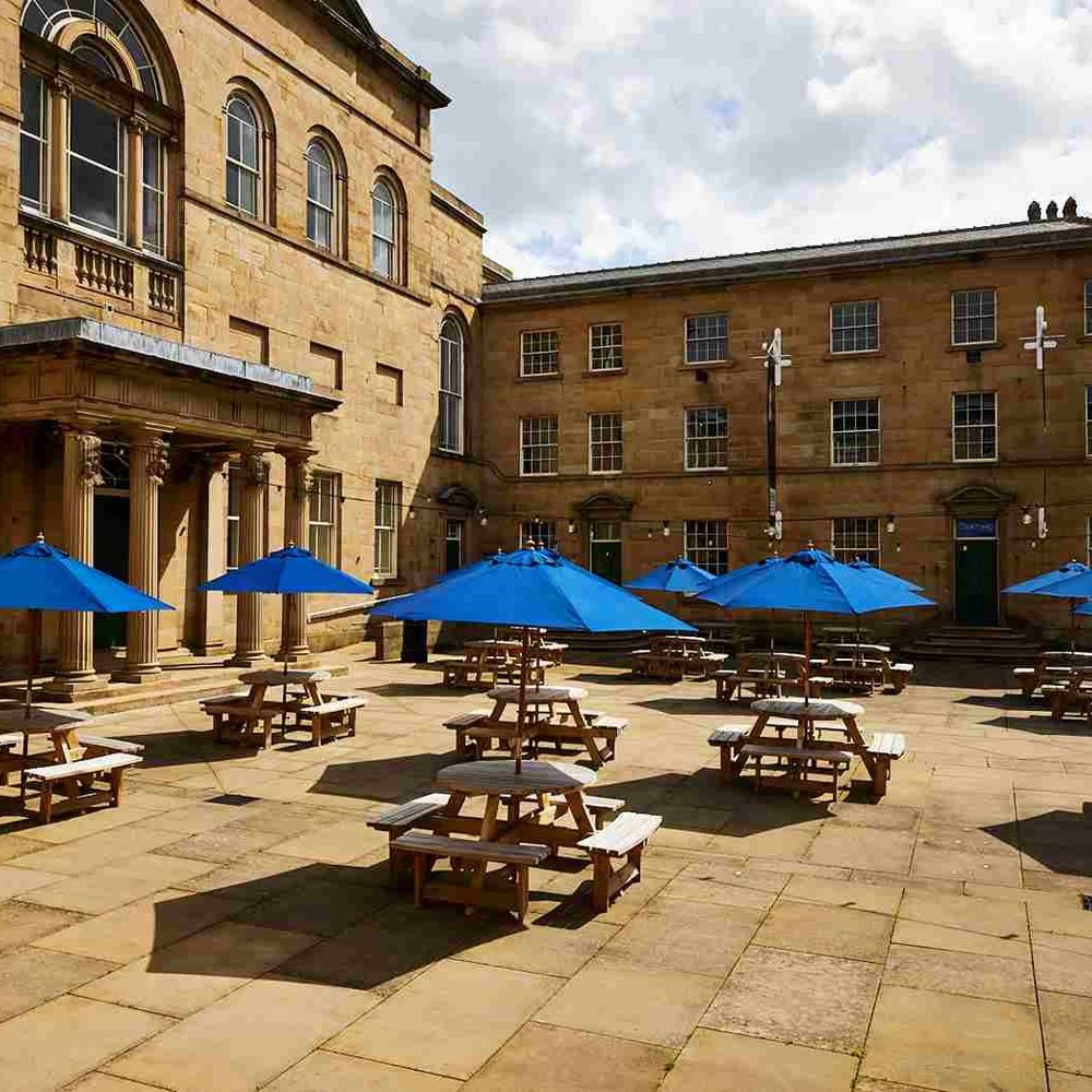 A courtyard with multiple round, wooden benches that each have blue umbrellas in. 