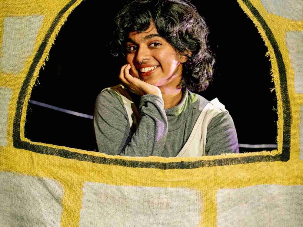 A performer smiles from a tower window, resting her chin on her hand. The yellow fabric tower, painted with brickwork, represents Rapunzel's prison in this colourful stage adaptation.