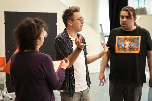 Three people engaged in a discussion during a rehearsal. The person in the middle, wearing glasses and gesturing with both hands, appears to be leading the conversation. A woman with curly hair and glasses holds a red folder, listening attentively. A man in a black graphic T-shirt stands to the right, holding a blue marker, also listening. The background shows a light-filled rehearsal space with a black curtain and white walls