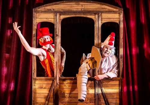 A clown hangs out of a wooden train window. Another character wearing a big, silly, red moustache stands beside the clown with his arm raised leaning out the window.