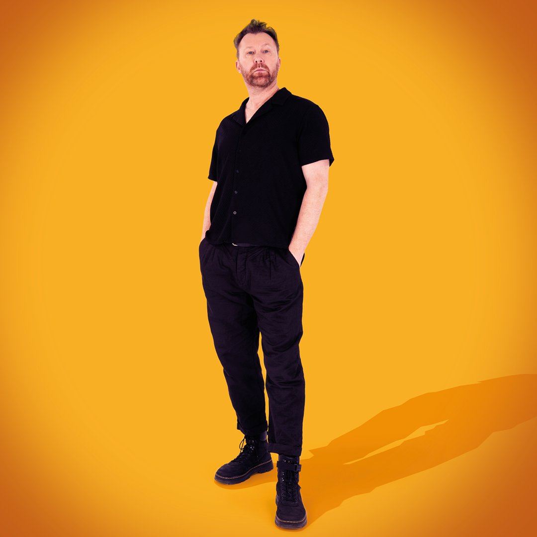 A man dressed in black stands with his hands in his pockets against a bright orange background, looking confidently at the camera.
