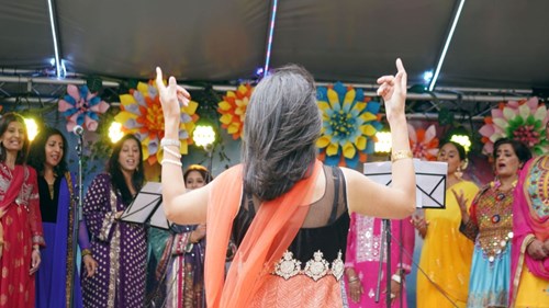 A group of performers dressed in colourful traditional South Asian attire sing on stage, led by a conductor whose back is to the camera.