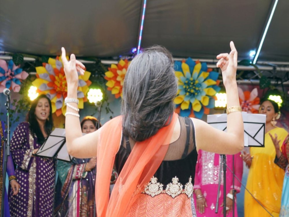 A group of performers dressed in colourful traditional South Asian attire sing on stage, led by a conductor whose back is to the camera.