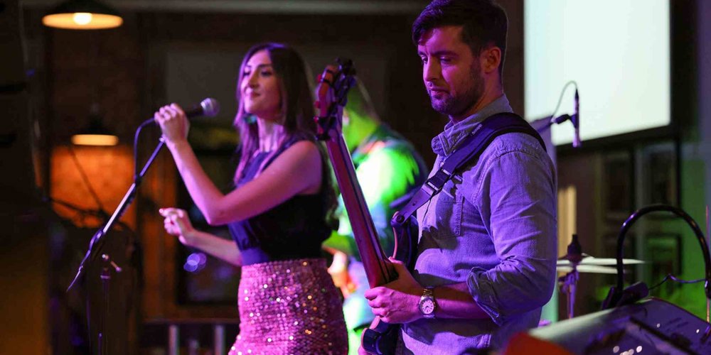 Female singer performs into a microphone while a guitarist plays beside her under colourful stage lighting at a live music event.
