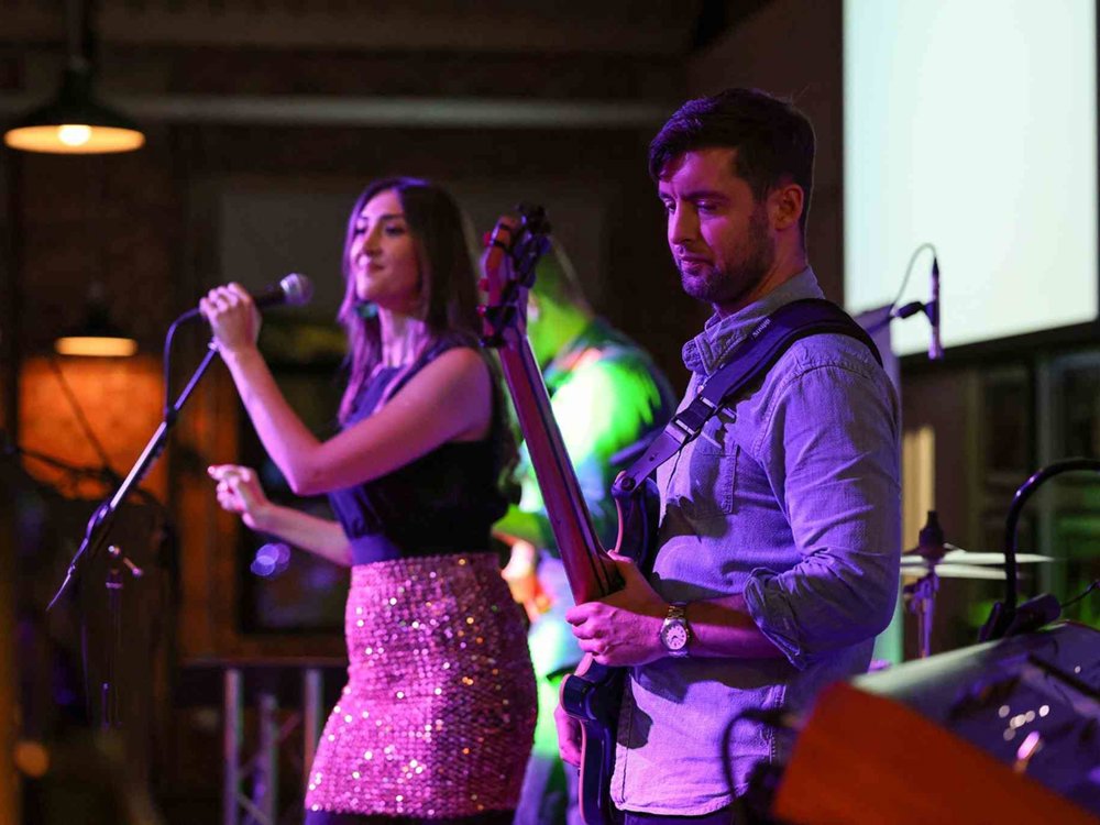 Female singer performs into a microphone while a guitarist plays beside her under colourful stage lighting at a live music event.