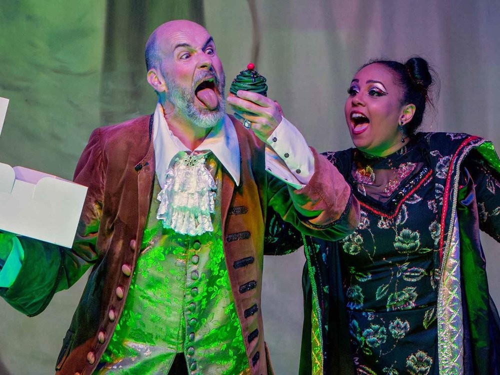 A comedic scene with a bearded man eagerly reaching toward a green cupcake while a woman beside him reacts in shock and delight under dramatic green lighting.