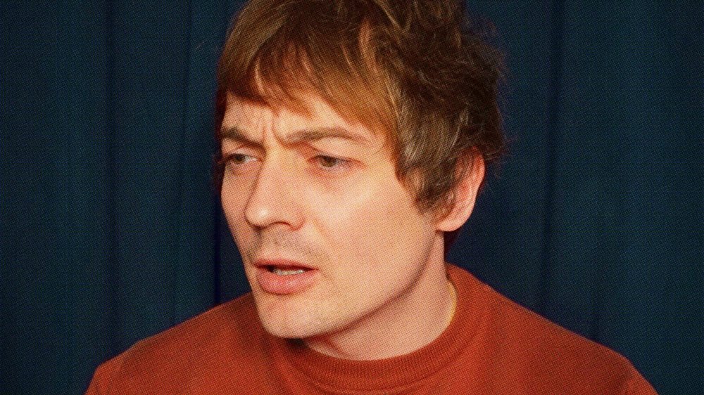 A close-up portrait of a person with short brown hair wearing a red top, looking slightly to the side with a thoughtful expression against a dark blue background.