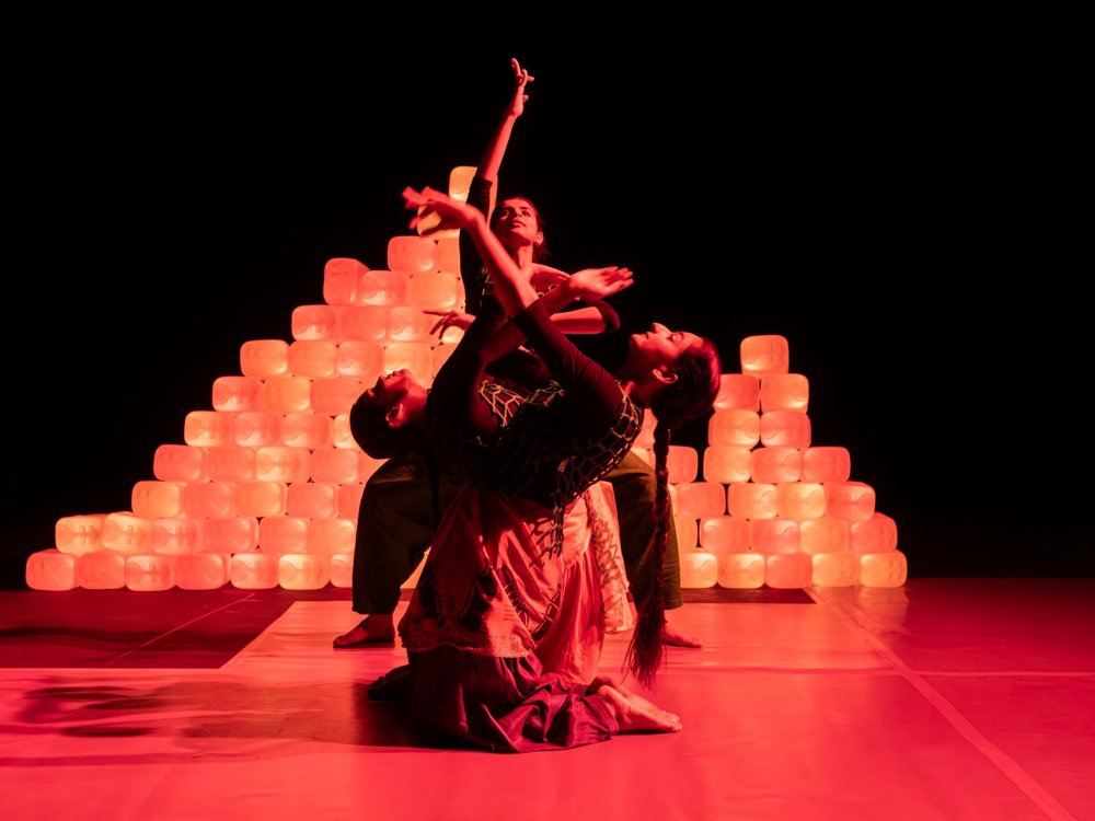 Three dancers create an interwoven sculptural pose under red lighting, framed by a glowing pyramid of stacked plastic containers behind them.