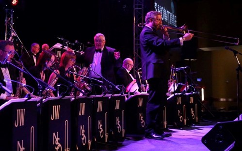Dressed smartly in a suit, a person stands in the middle of the stage playing a trombone. Behind them, the rest of the orchestra play various musical instruments.