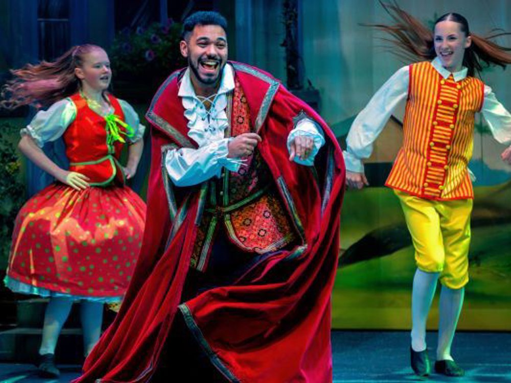 Three pantomime performers dance energetically on stage, led by a smiling man in a red cape, flanked by two performers in colourful storybook costumes, mid-movement under bright stage lighting.
