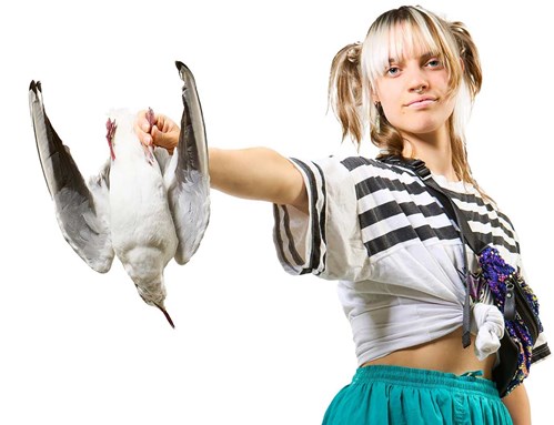 A young person with blond and brown hair has her arm outstretched, holding a seagull. She is wearing light blue trousers and a black and white striped tshirt tied up with a bag across her chest. 