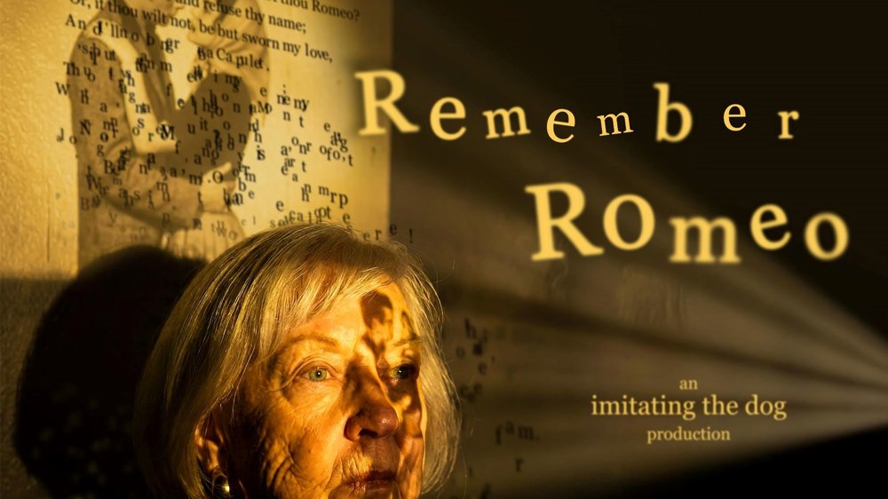 Promotional image for Remember Romeo. An older woman with silver hair is illuminated by warm golden light as projected text and images from Romeo and Juliet fall across her face and the wall behind her. The shadow of a young figure appears over a page of Shakespeare's text, while the title "Remember Romeo" is projected prominently to the right.