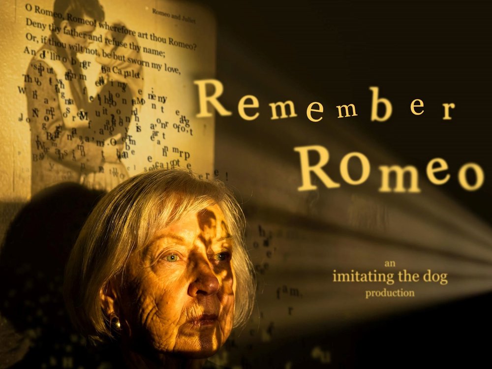 Promotional image for Remember Romeo. An older woman with silver hair is illuminated by warm golden light as projected text and images from Romeo and Juliet fall across her face and the wall behind her. The shadow of a young figure appears over a page of Shakespeare's text, while the title "Remember Romeo" is projected prominently to the right.