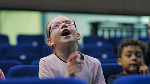 A young girl with light skin, wearing pink glasses and a light pink t-shirt, sits in a theatre with blue seats. She is looking up with an excited expression, her mouth open and her hand raised in a gesture. Behind her, a young boy with curly hair and darker skin sits in a similar blue seat, rubbing one of his eyes with his finger. The background is slightly blurred, showing additional rows of blue theatre seats.