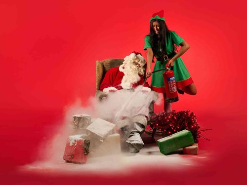 A comedic Christmas scene with Santa slumped in a chair, covered in white fire extinguisher foam, while a playful elf in a green costume sprays him with a red fire extinguisher. Burnt and scorched presents surround them, set against a bright red background.
