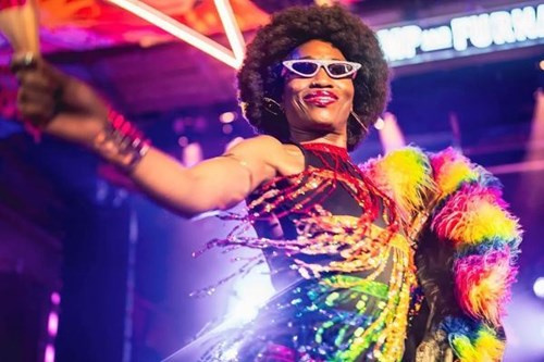 A vibrant performer on stage wearing a sparkling, multicoloured outfit with fringe details and a rainbow feathered boa. They have an afro wig, cat-eye sunglasses, and bold makeup, exuding confidence and joy while extending their arm in an expressive pose. The background is illuminated with bright lights and neon signage, suggesting a lively event or nightclub setting.