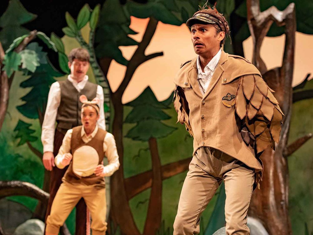 A bird-like character in layered, feather-inspired clothing stands in the foreground looking worried, while two smaller animal characters react behind him on a forest stage.