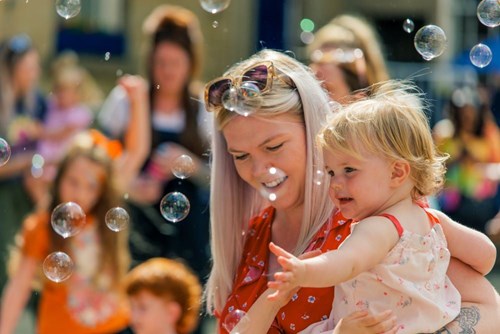 Outside on a sunny day, a woman with blonde hair is holding a toddler up, the toddler is playing with bubbles in the air