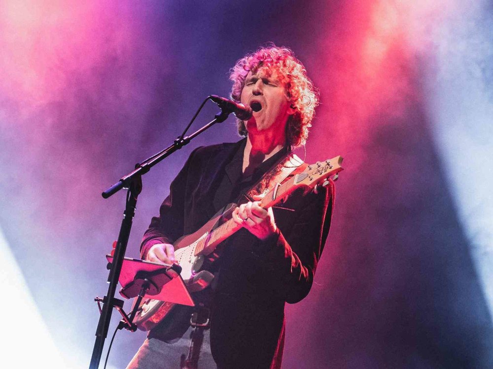 A musician on stage sings passionately into a microphone while playing an electric guitar, illuminated by strong pink and blue stage lighting with drifting smoke behind them.
