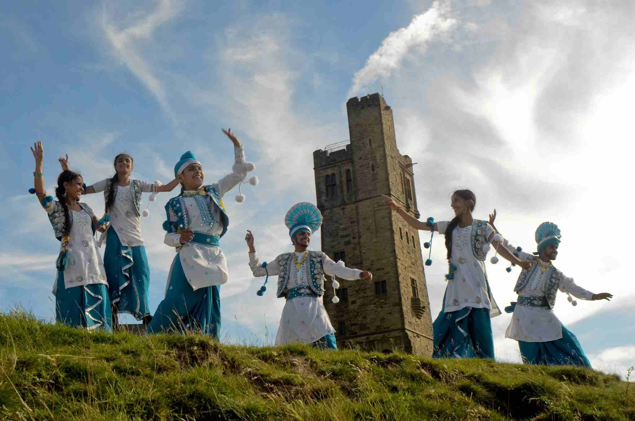 Virsa Bhangra dancers in blue and white traditional costumes perform outdoors on Castle Hill, with Victoria Tower rising behind them beneath a bright blue sky.