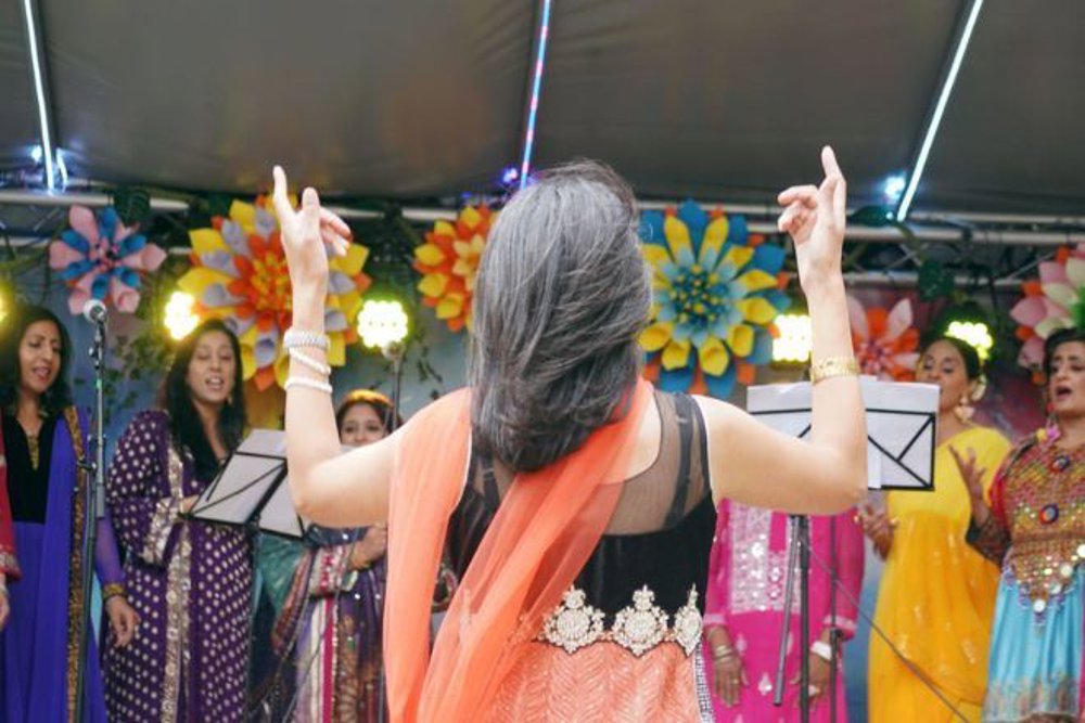 A group of singers dressed in vibrant traditional attire performing on an outdoor stage, directed by a conductor wearing an orange and black outfit with her back to the camera. The stage is adorned with colourful floral decorations and bright lights.