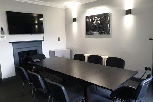 Small Meeting Room at Lawrence Batley Theatre with a large table with 8 chairs around. The walls are a light grey and with a photograph on one wall and a TV on another.