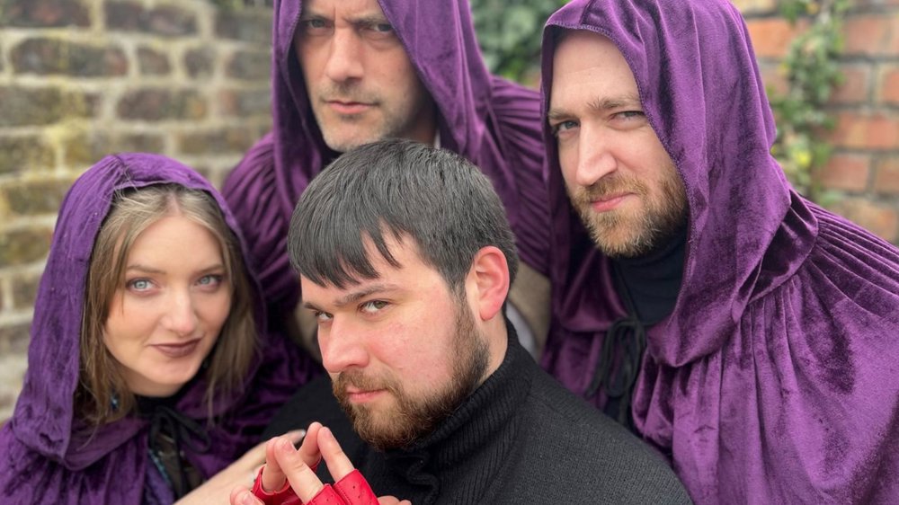 Four people pose closely together outdoors against a brick wall. Three wear deep purple hooded cloaks and look intensely at the camera, while a fourth person in a black jumper and bright red fingerless gloves sits in front with their hands clasped. The group’s dramatic expressions and costumes create a mysterious, fantasy-inspired atmosphere.