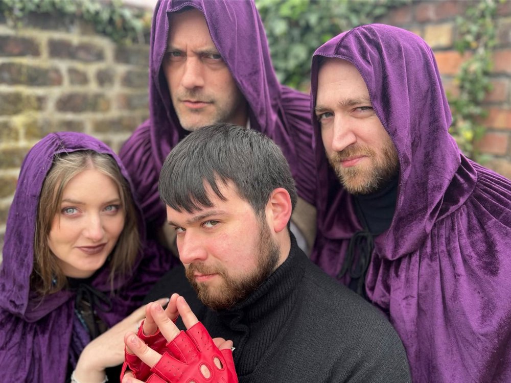 Four people pose closely together outdoors against a brick wall. Three wear deep purple hooded cloaks and look intensely at the camera, while a fourth person in a black jumper and bright red fingerless gloves sits in front with their hands clasped. The group’s dramatic expressions and costumes create a mysterious, fantasy-inspired atmosphere.