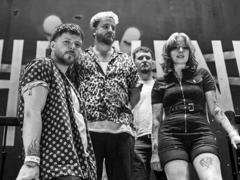 Four musicians pose together in a black and white photo, standing confidently on a staircase and looking down at the camera.