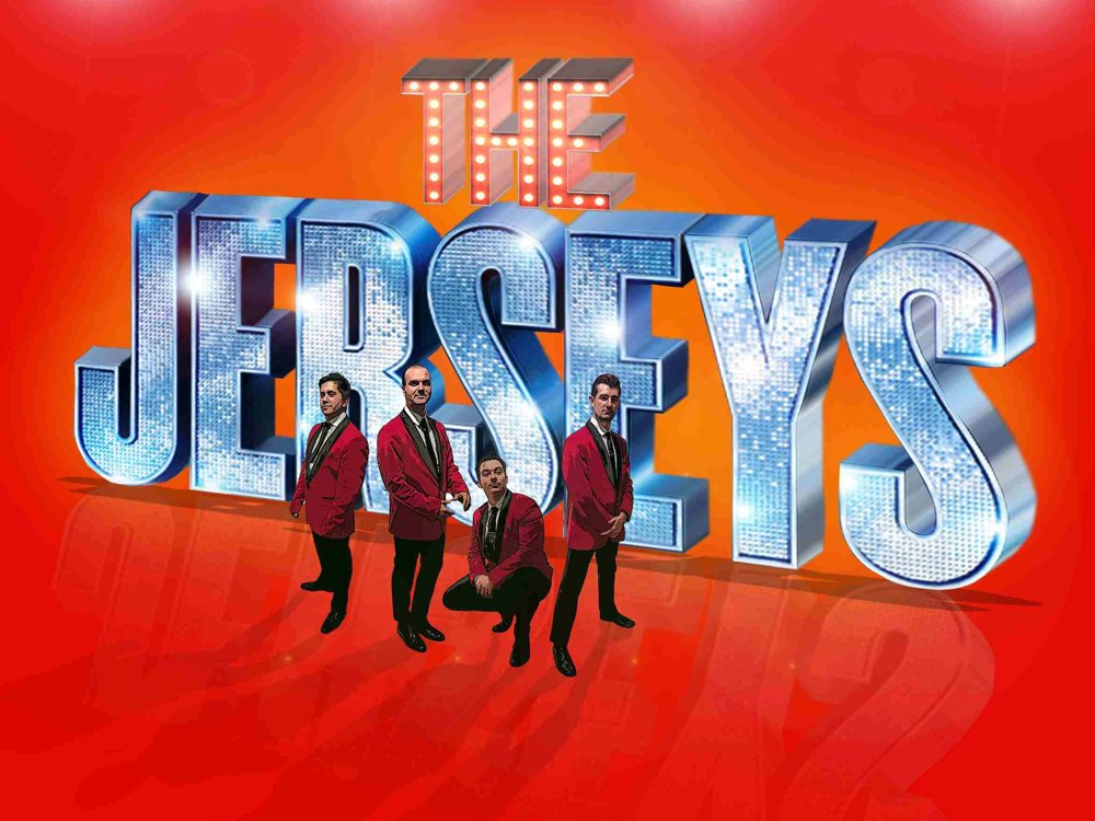 Four men in matching red blazers pose confidently in front of giant sparkling 3D letters spelling “The Jersey Boys” on a bright red and orange background with glowing stage lights overhead.