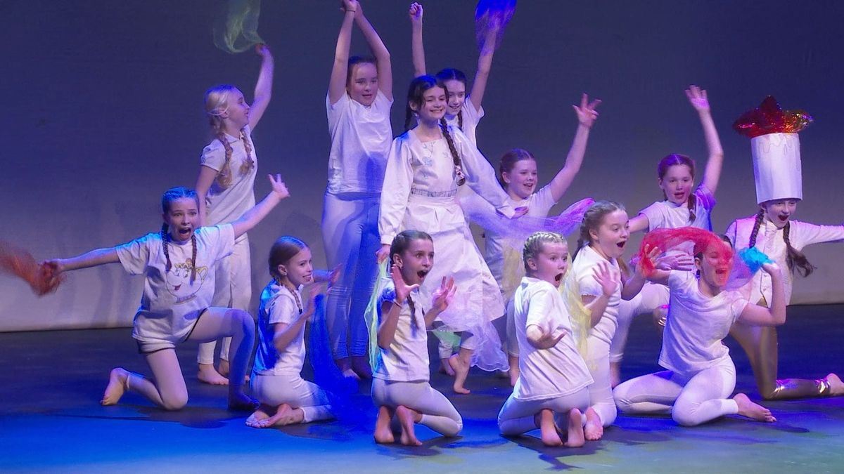 A group of children in white costumes perform an energetic dance routine on a theatre stage, striking playful poses under blue and purple stage lighting.