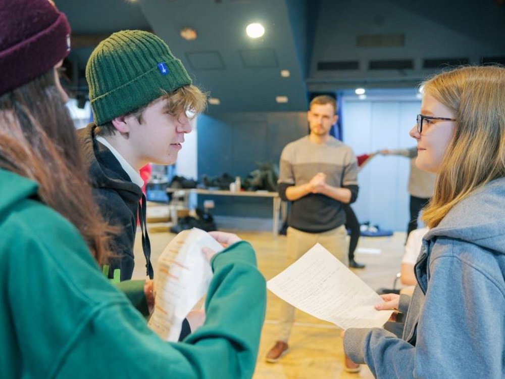 A group of young people engaged in a theatre rehearsal in a spacious, well-lit room. Three individuals in the foreground, dressed in hoodies and beanies, are holding scripts and conversing intently. One of them, a young woman with glasses in a grey hoodie, faces a young man wearing a green beanie, while another person in a maroon beanie listens. In the background, a facilitator or director in a two-toned jumper observes with hands clasped, while other participants are engaged in activities.